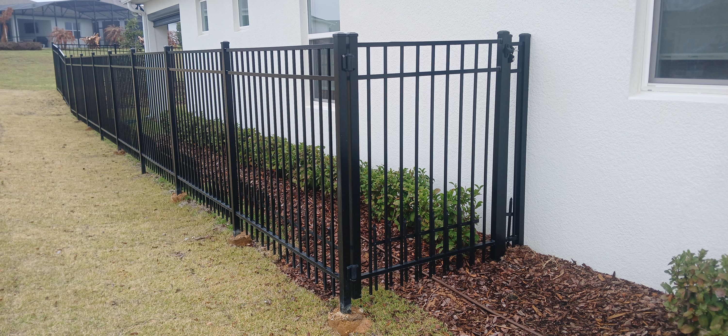A black powder-coated aluminium picket fence and gate beside a house, set over a fresh mulch bed