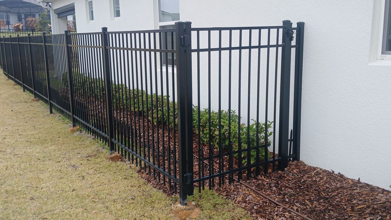 A black powder-coated aluminium picket fence and gate beside a house, set over a fresh mulch bed