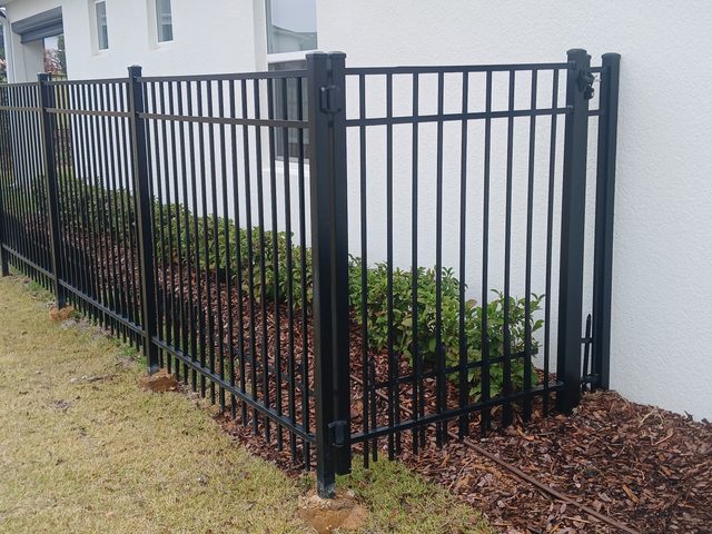 A black powder-coated aluminium picket fence and gate beside a house, set over a fresh mulch bed