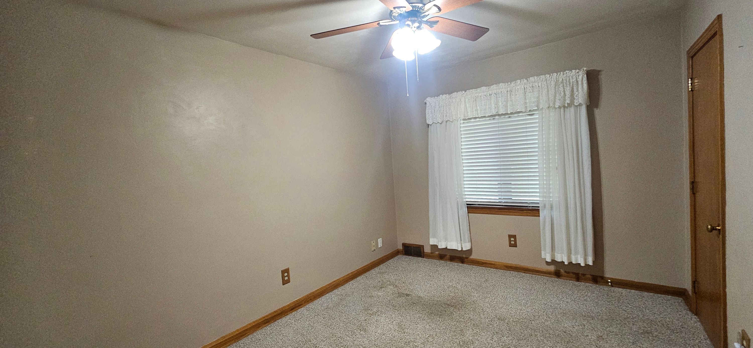 Bedroom before painting with tan walls, white curtains and a ceiling fan