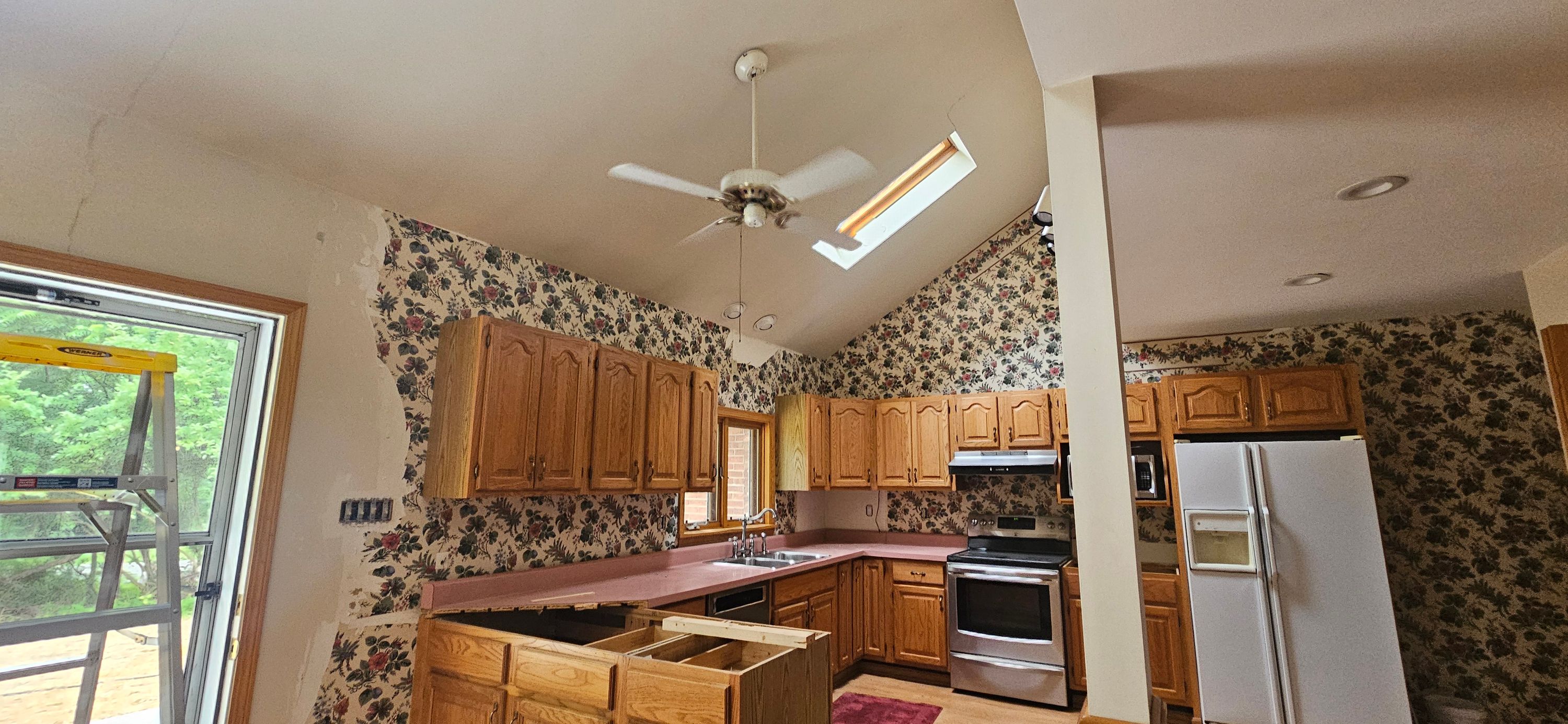 Kitchen before work with dated floral wallpaper covering the walls above oak cabinets