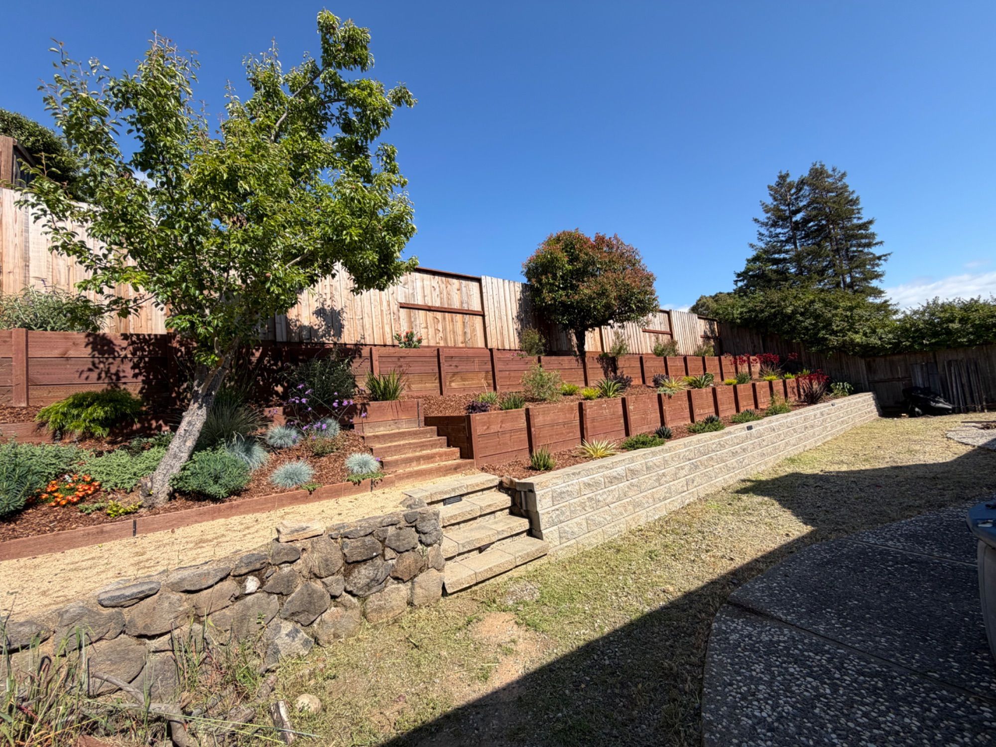 A terraced hillside garden held by timber retaining walls above a river-rock wall, planted and mulched