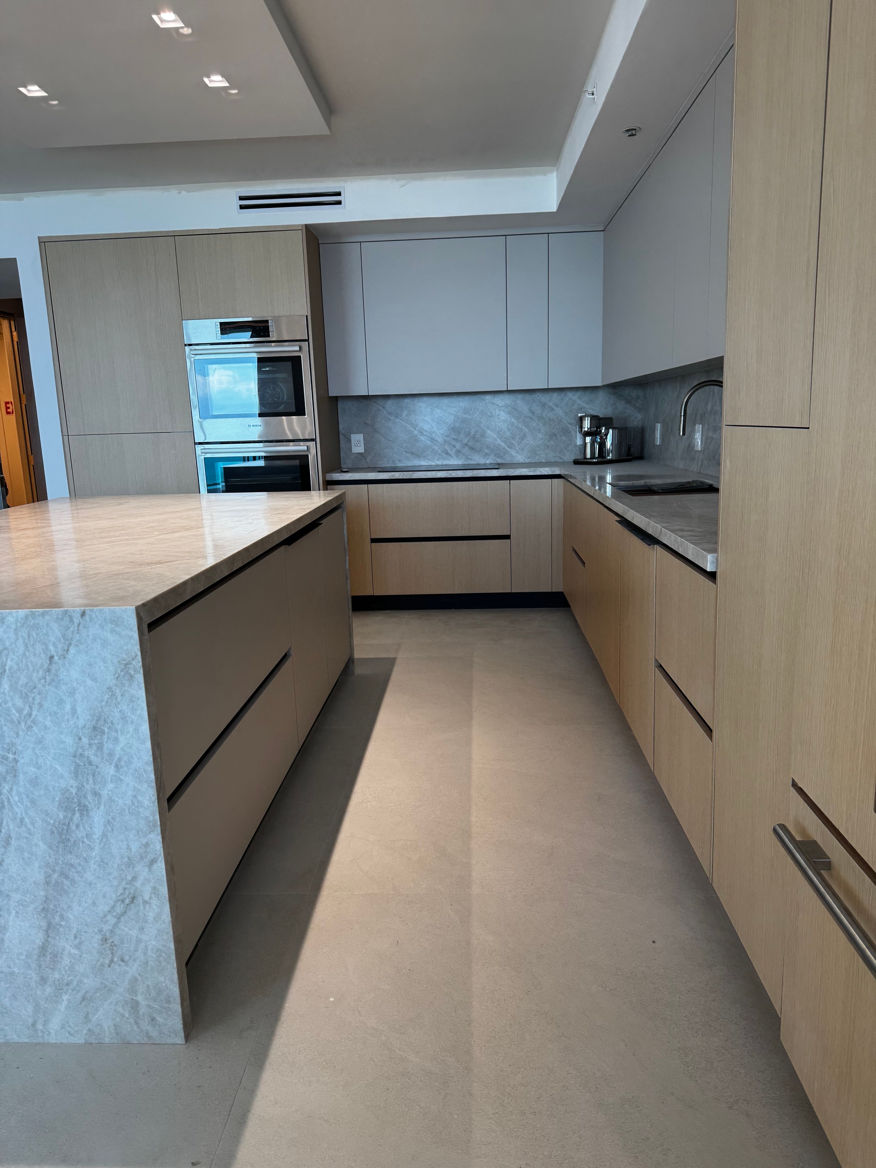 Light oak handleless kitchen with marble waterfall island, double wall ovens, and stone backsplash