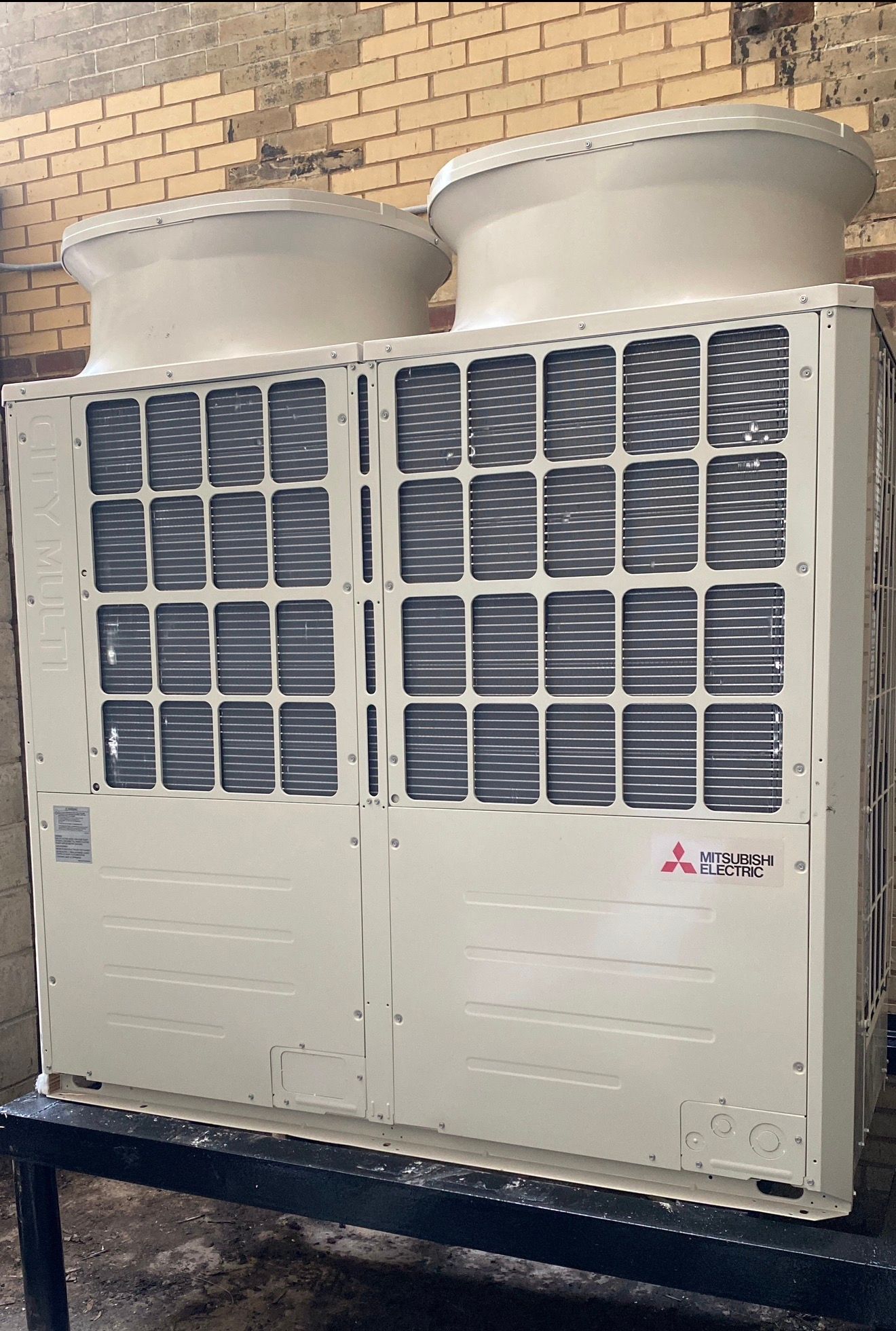 Two outdoor condenser units mounted side by side on a raised steel stand against a brick wall