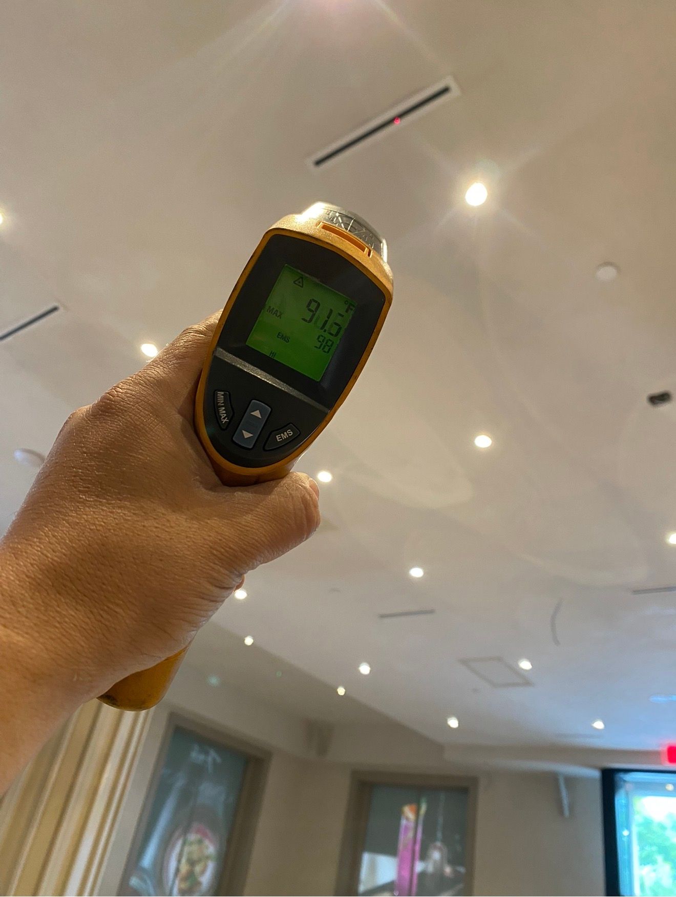 An infrared thermometer held up to a ceiling of linear slot diffusers to check supply air temperature
